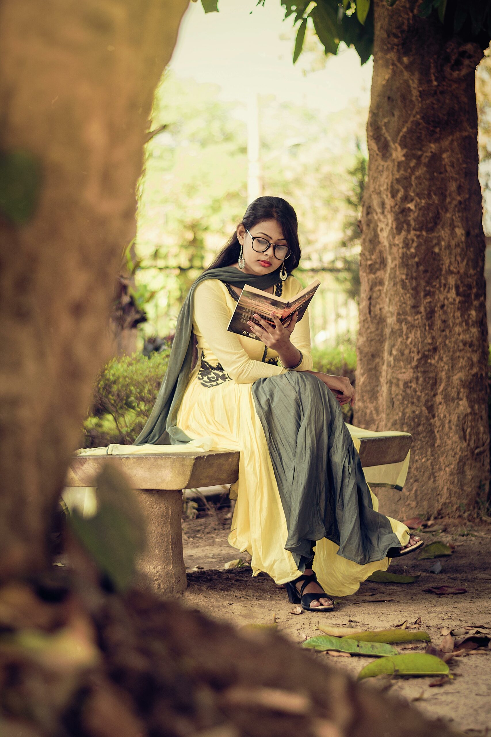 South Asian woman reading a book on a bench in a serene outdoor park setting.